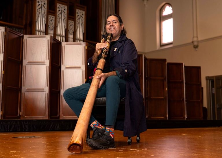 William Barton sitting on stage holding a painted yidaki (didgeridoo) in a hall with wooden paneling and a pipe organ.